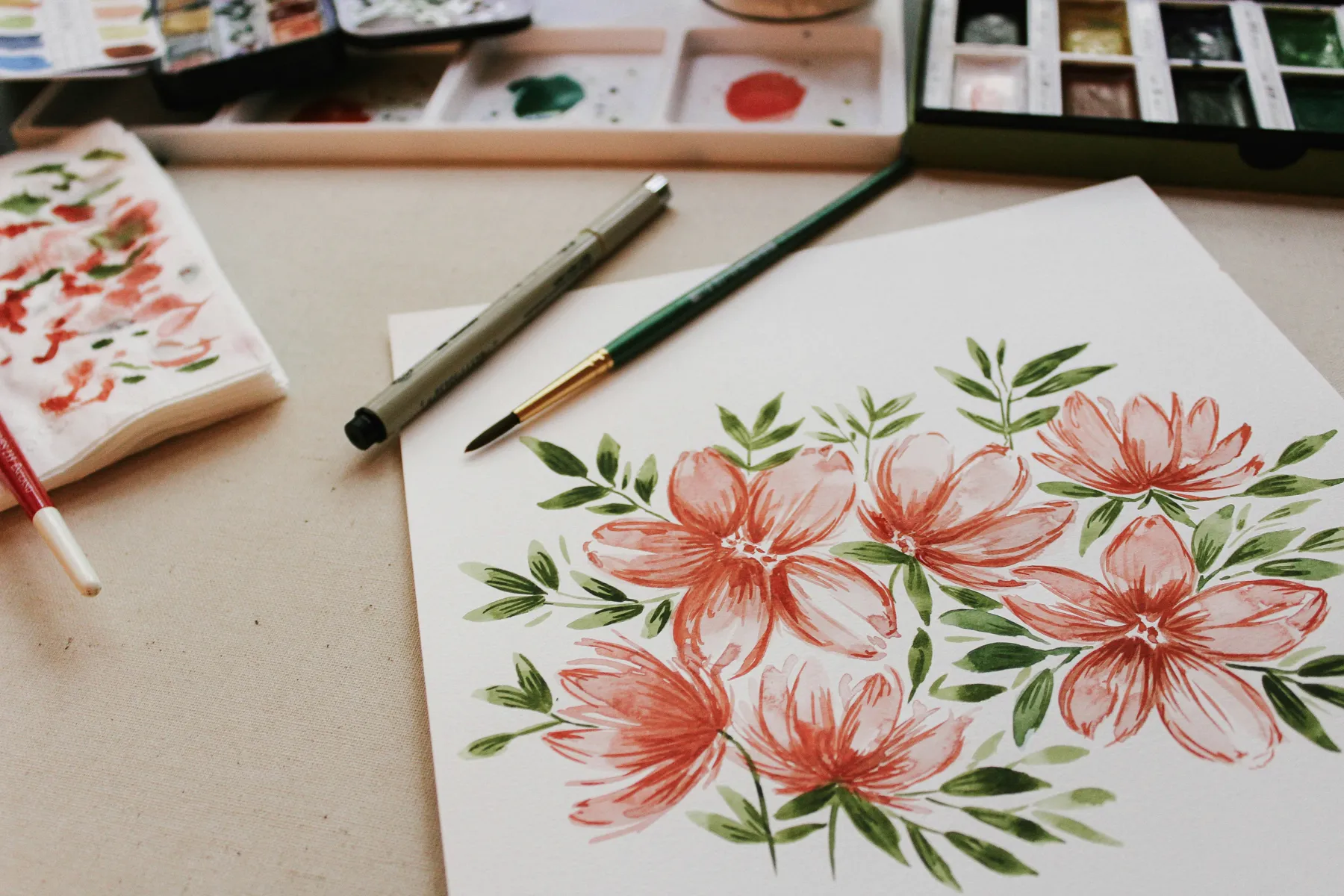 Coral watercolour flowers with green leaves on paper, beside a brush, a pen and a paint palette